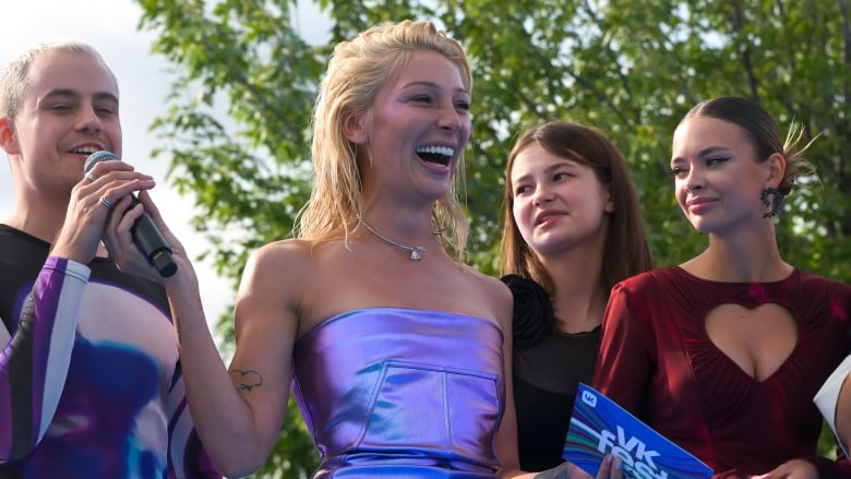 A blond woman wearing a blue dress is shown smiling during an outdoor event as she hands a microphone to a person behind her.