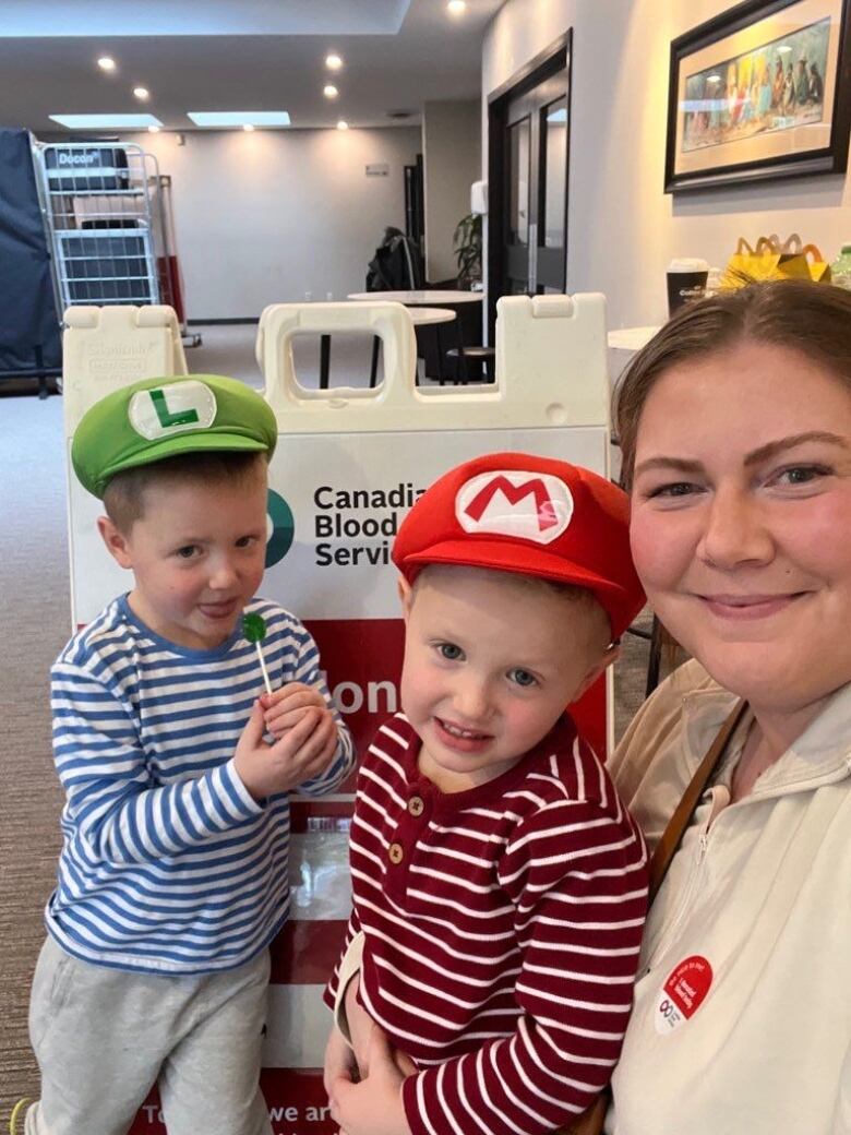 A woman takes a selfie crouched next to a sign for Canadian Blood Services, with two young sons dressed as Mario and Luigi characters next to her.