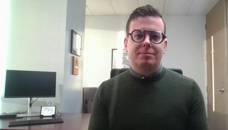 A man wearing a sweater and glasses sits at an office desk for an interview via video-chat software.