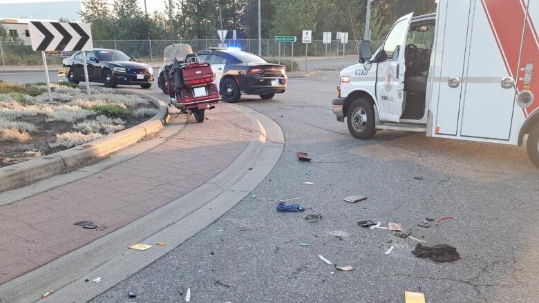 A motorcycle is pictured on the road with police cars and an ambulance. Various items and parts are strewn on the ground. 