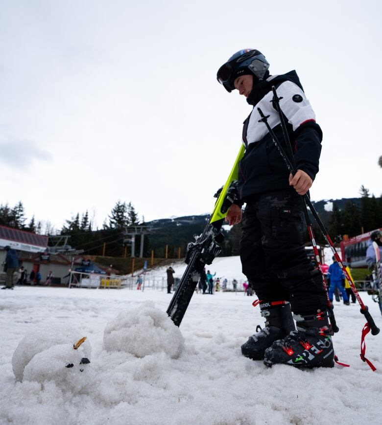 A person in ski clothes stands next to a melted snowman with a mountain behind them.