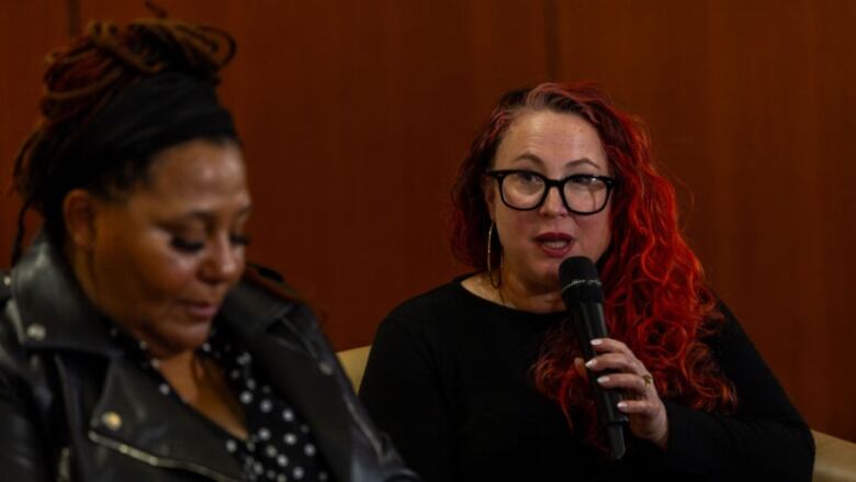 A woman sits holding a microphone speaking on a panel, with another woman's profile in the foreground.