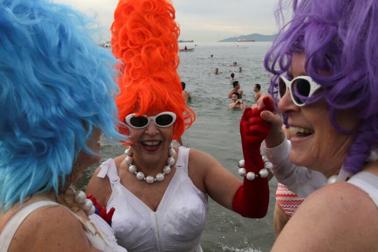 Three women wearing elaborate coloured wigs and glam-era costumes smile while partially submerged in beachwater. Others are visible behind them.