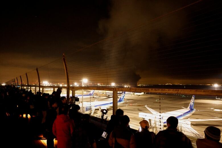 People line up against a fence at an airport as a large black cloud floats skyward in the distance.