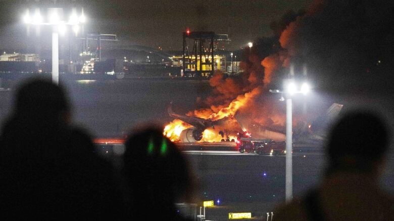 Silhouette of three people in the foreground watching a plane engulfed by fire on the tarmac at night.