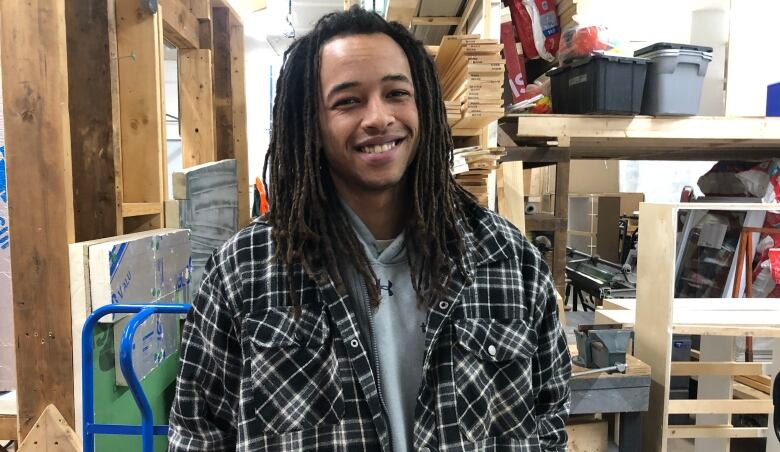 A man with dreadlocks in a plaid shirt stands in front of a wooden frame and wood planks in a warehouse.