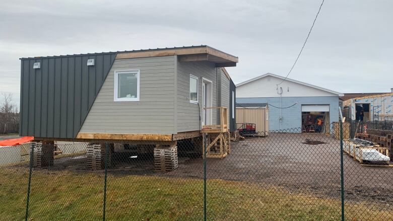 A small building rests on pallets in a parking lot. It has black and beige siding and small windows, with a staircase up to the front door.