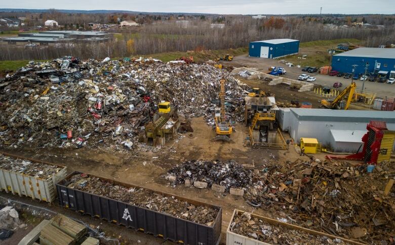 Several large rail cars filled with various items. Heavy equipment in the background is surrounded by piles of scrap metal and several buildings.