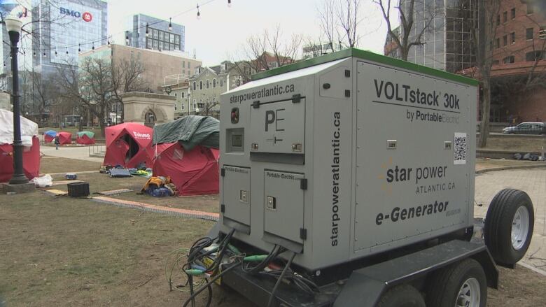 A large white generator with a green top sits on a wheeled stand. In the background, there are tents.