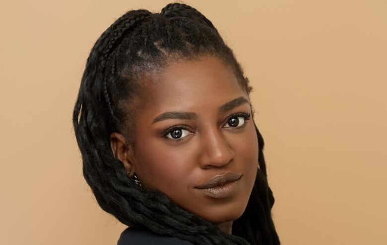 A young Black woman with long braided hair looks at the camera in front of a plain tan background. 