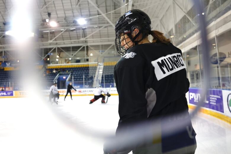 A hockey player is on the ice with a helmet and a ponytail. She's by the boards with hockey action in the background.