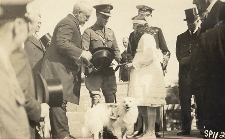 A black-and-white picture of a prince receiving a postcard from a girl, with a dog in the foreground. Other officials are around them.