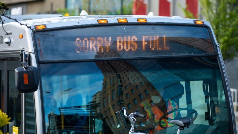 A bus driver wearing a high-vis vest and a mask waits at a stop, with a sign above him reading 'Sorry Bus Full'.