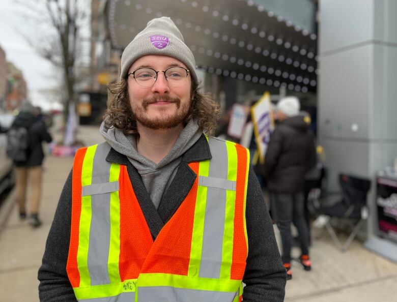 A man with glasses and a beard wears a touque and safety vest while standing on a sidewalk.