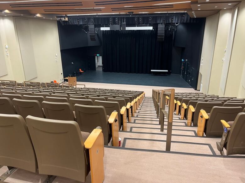 A lecture theatre with folding seats facing a large stage with a microphoned podium and a black stage floor. 