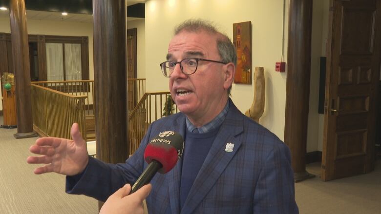 A man with glasses in a dark blue suit speaks outside city council chambers.