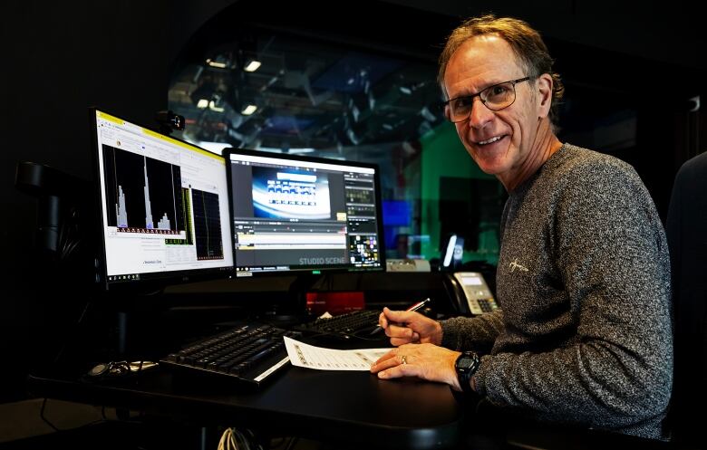 A man sits in front of two computer monitors but smiles at the camera