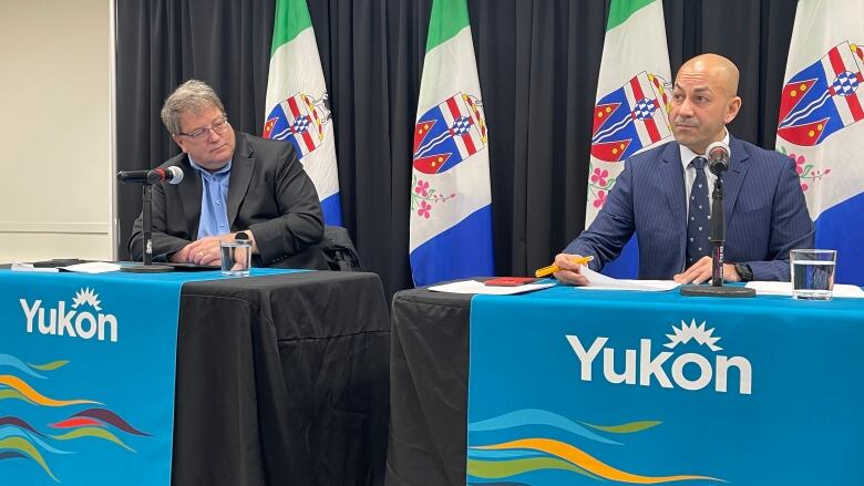 Two men sit behind tables with microphones and 'Yukon' banners.