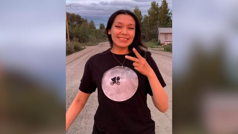 A young person stands outside and smiles while giving the 'peace sign' with their fingers.