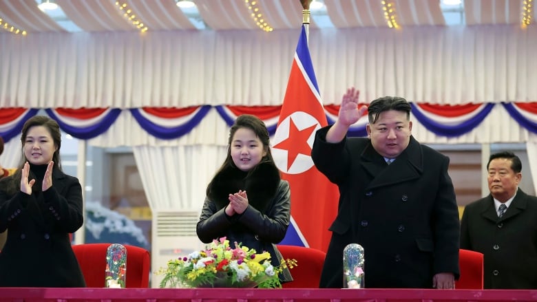 Kim Jong-un waves as his family members clap at a table in front of the North Korea flag.