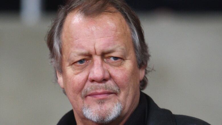 An older man in a black shirt and jacket slightly scowls as he looks to his left.