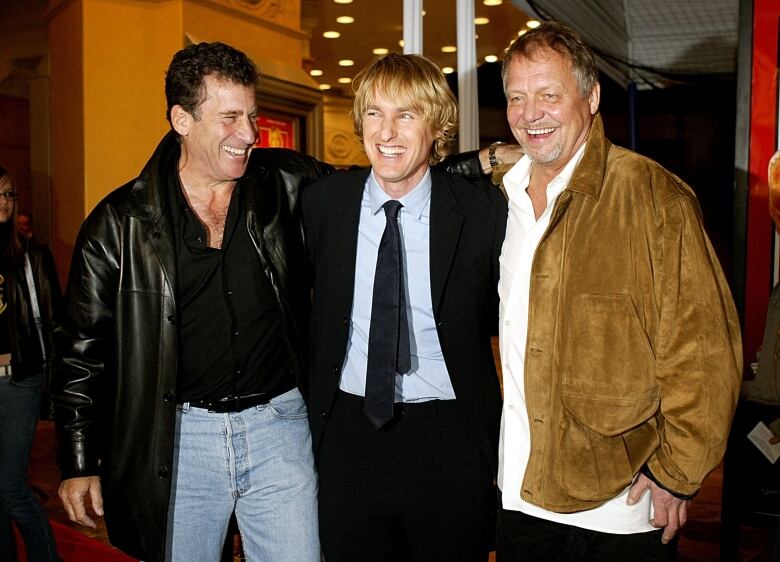 Three men standing in a line with their arms around each other smile broadly outside of a movie theatre.