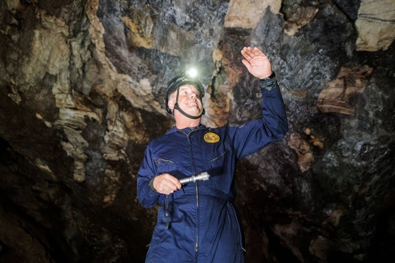 The scientist stands in blue coveralls with a helmet and light on his head deep inside a cave system.