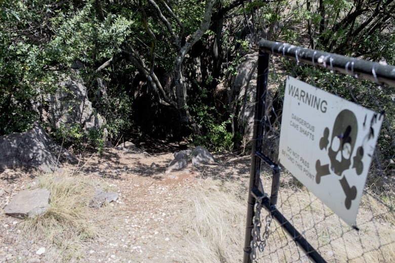 A dark hole in amongst trees and shrubs are gated off with a warning sign to not enter is the entrance to the cave system.