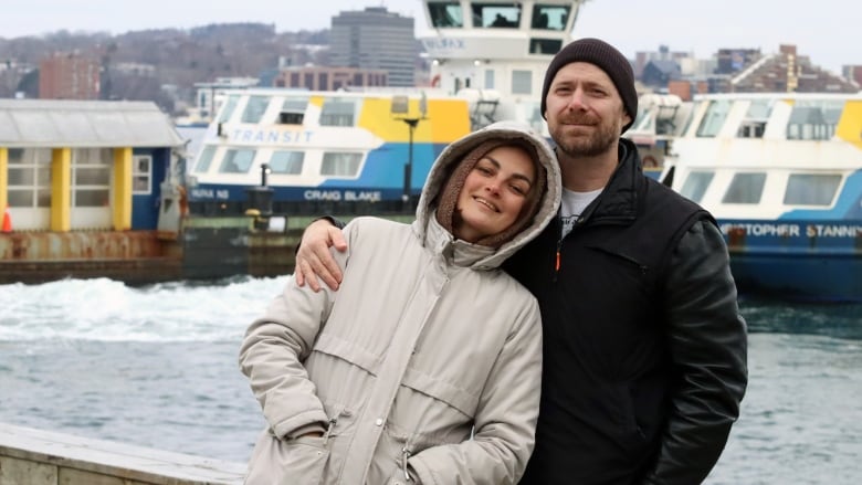 A man in a black coat puts his arm around a woman in a grey coat. A ferry can be seen in the water behind them.