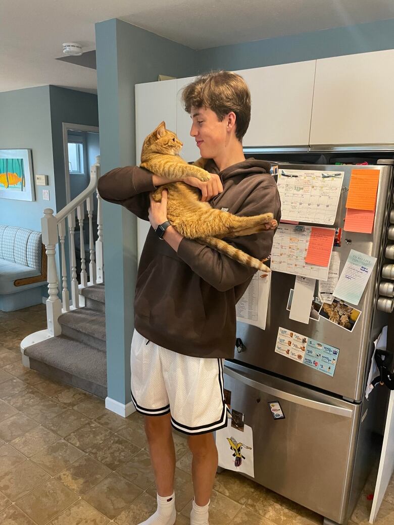 A teenage boy holds an orange cat in his hand and looks closely at it next to a fridge.