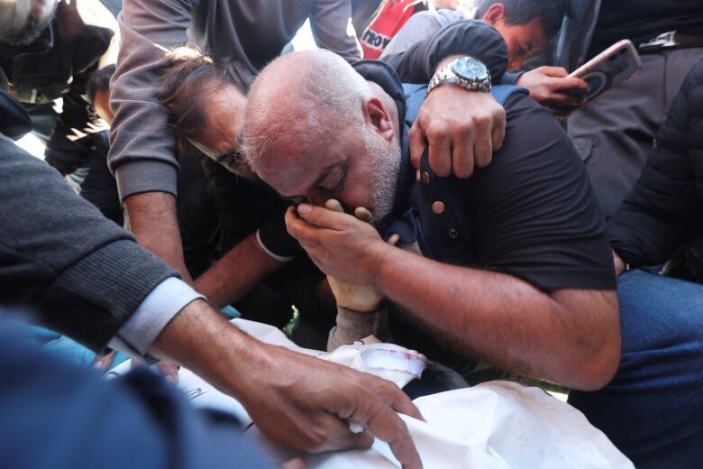 A mourner covers their face and crouches over a body as another person embraces them.