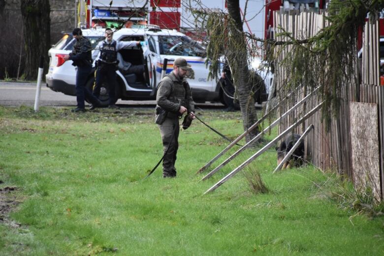 A police dog handler has a dog on a leash, which is sniffing a long stretch of wooden fence with emergency vehicles in the background.