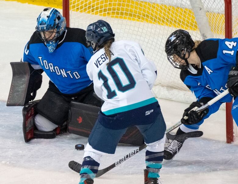 Three hockey players, two in blue and one in white, scramble around the net. 