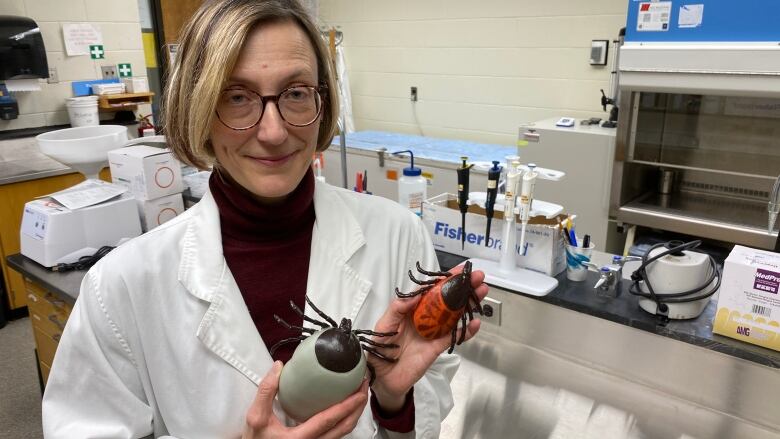 A woman wearing a lab coat can be seen holding two fake ticks in her hands. 