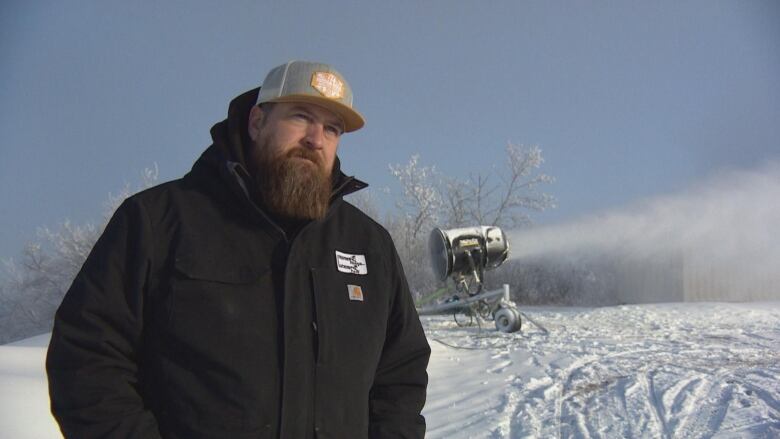 A man in a cap and a black jacket stands in from of a snow machine on a ski hill. 