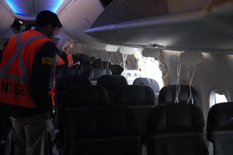 Two people wearing bright neon orange vest stand inside a passenger plane looking at a hole left behind after a panel ripped off. 