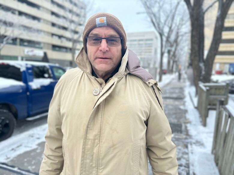 A man in a brown toque and beige winter coat stands on a snowy sidewalk in front of cars. 