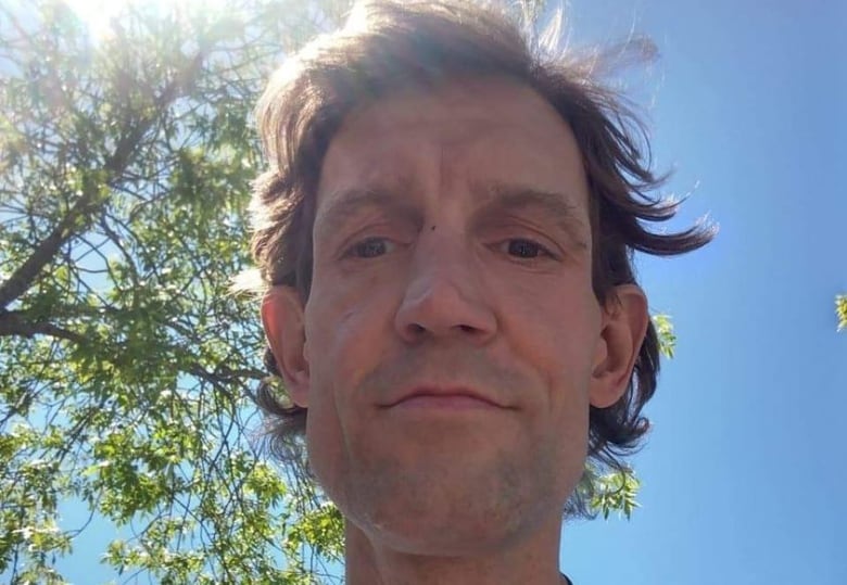 A selfie of a man wearing a white shirt and backpack with the blue sky, sun and tree in the background.