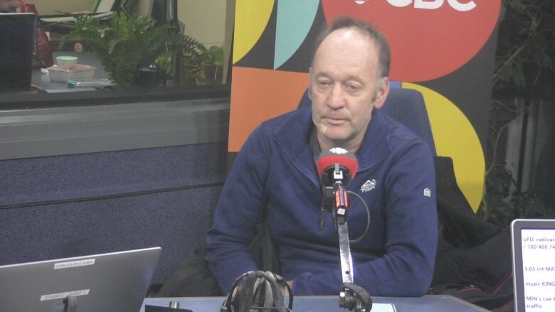 An older man with short brown hair sits in front of a microphone during an interview.