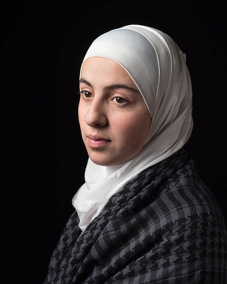 A woman cloaked in a grey scarf wears a white hijab.