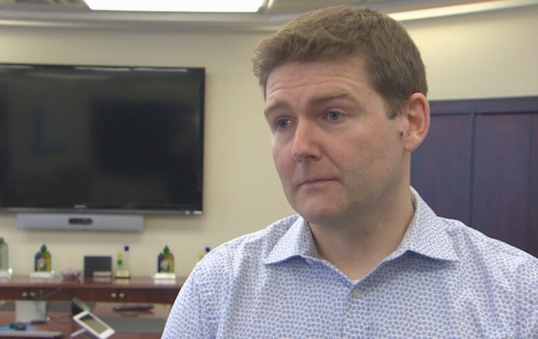 A shot from the chest up of a man, looking just off of the camera, in an office. There is a television and a desk behind him. 