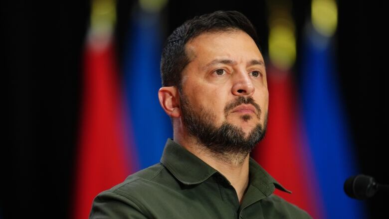 A man in an olive green shirt stands in front of a microphone. A row of Canadian and Ukrainian flags hang in the background.