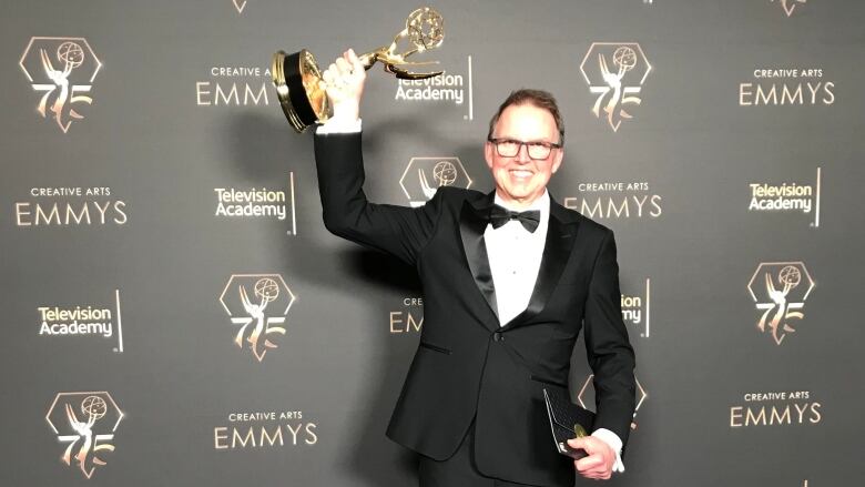 A man stands in front of a black patterned background holding a gold statue. 