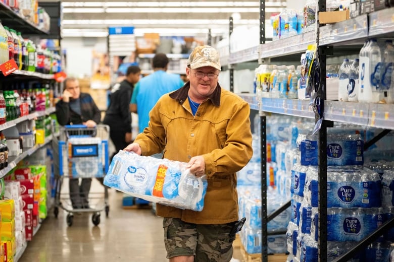 A man carries a case of bottled water in a supermarket aisle.