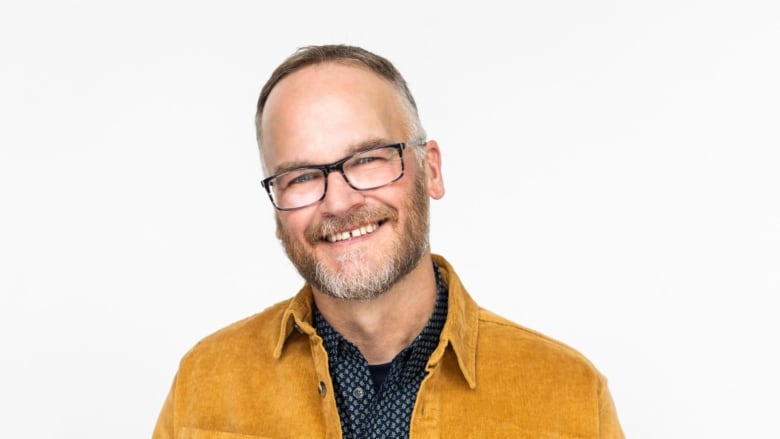 Man in came colour jacket and tshirt wearing glasses and a beard smiles to camera