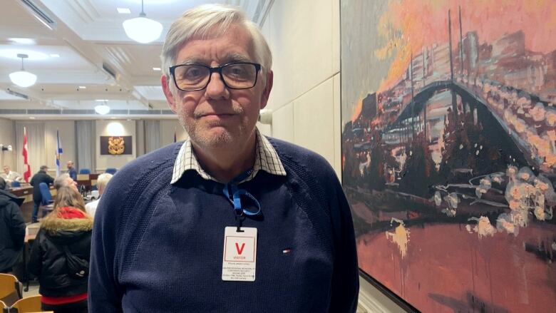 A white man with glasses and white hair stands in an open room with official Halifax city crest on the wall in the distance 