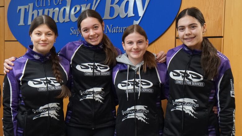 Four young people stand together and smile indoors. They are wearing purple and black jackets.