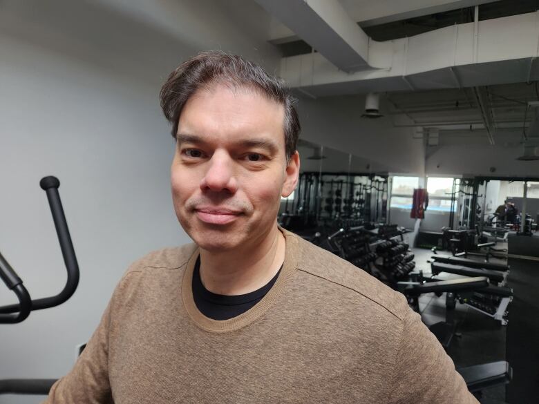 A man in a brown sweater inside a non-descript gym looks into the camera.