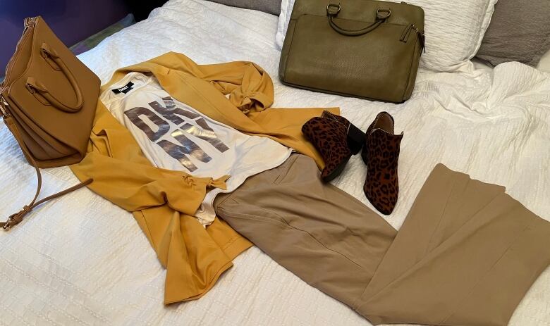 A womans outfit, including designer handbags, shoes, pants, jacket and a T-shirt with DKNY spelled in large letters, is arranged on a bedspread.