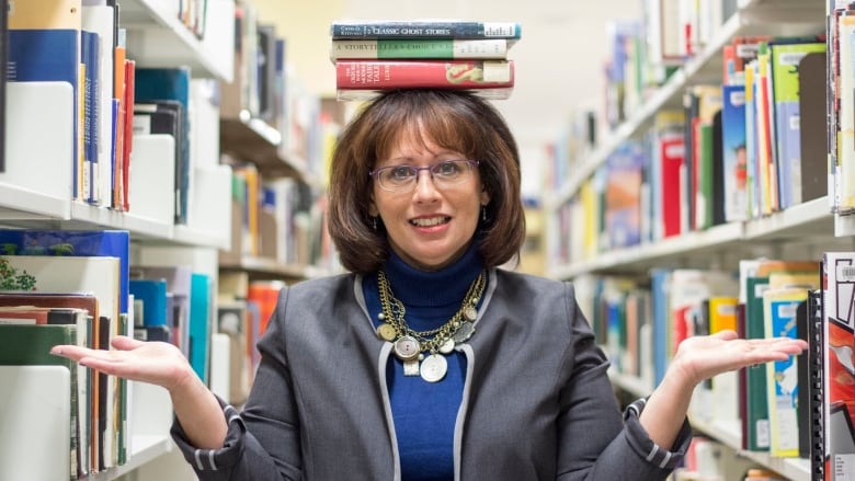 A woman balances three books on her head while gesturing to the shelves stacked with books on either side.  